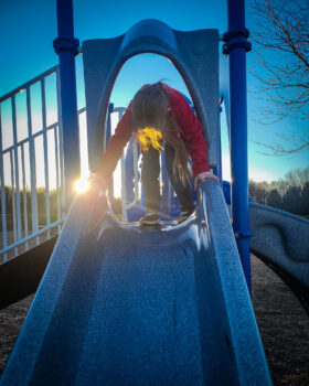 Kid at top of slide on playground, sun going down behind him.