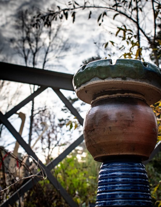 pottery sculputer, circular cone base, globe like middle with a bowl or dish atop, in front of a fence with dramatic cloudy sky behind